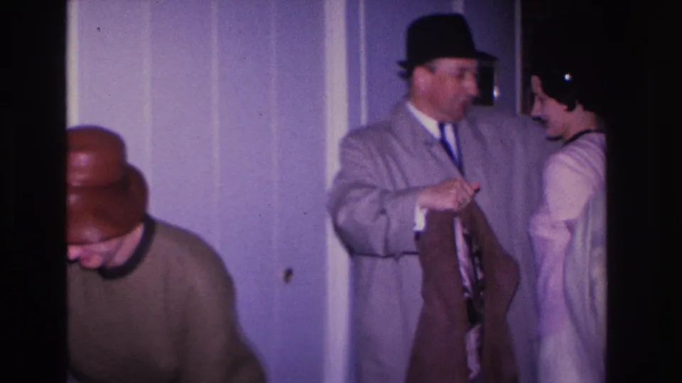Man in a gray suit and dark fedora holds a coat near a woman in a light dress; an older adult wearing glasses is visible in the foreground, private interior circa 1966.