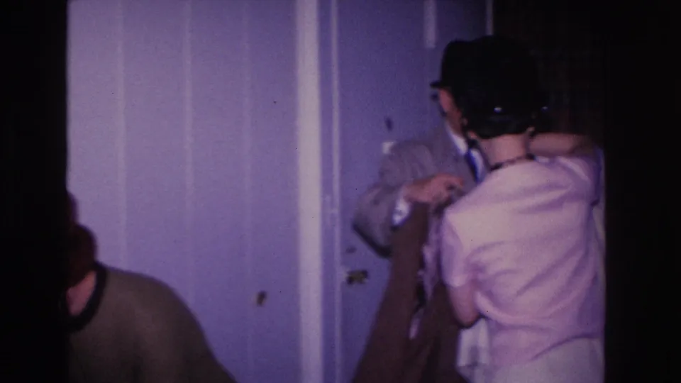 Small group of adults near a light blue painted interior wall, one man wearing a dark fedora and one woman in a light dress, private setting circa 1966.