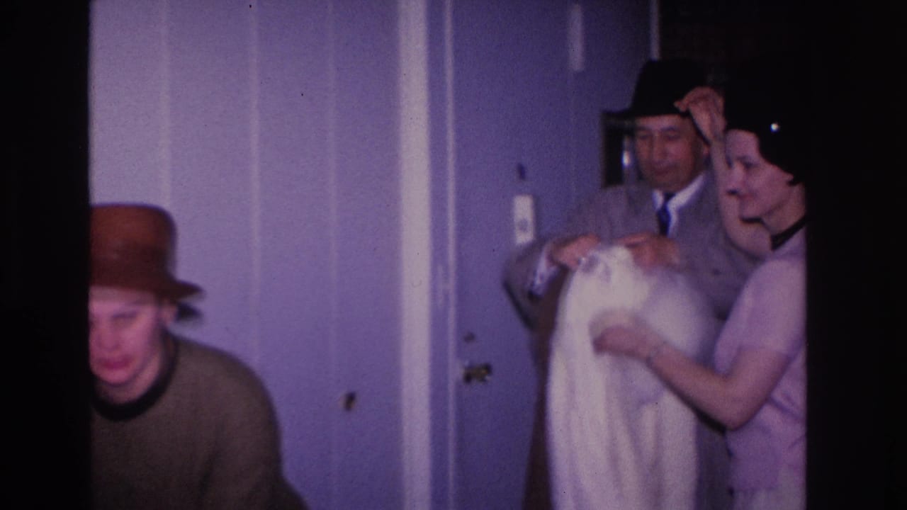 Three adults in a 1966 Hoboken, New Jersey residential hallway; man in gray overcoat and dark brim hat assists a woman in a pink dress while a second woman in a brown hat looks on.