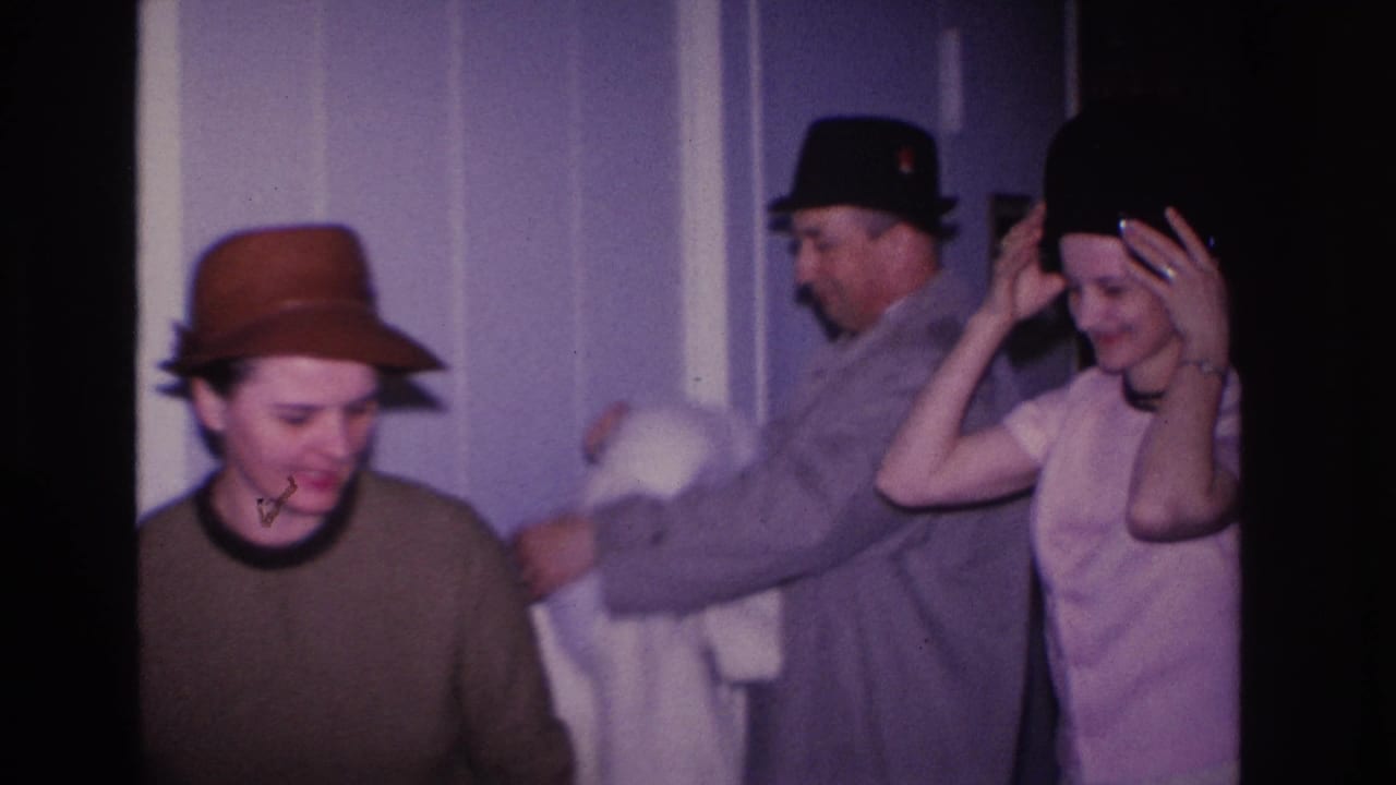 Three adults in a residential interior circa 1966 Hoboken, New Jersey; woman in brown brim hat looks toward camera while man in dark hat and gray coat assists second woman in pink dress.
