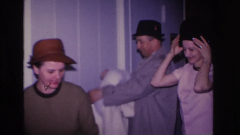 Three adults in a residential interior circa 1966 Hoboken, New Jersey; woman in brown brim hat looks toward camera while man in dark hat and gray coat assists second woman in pink dress.