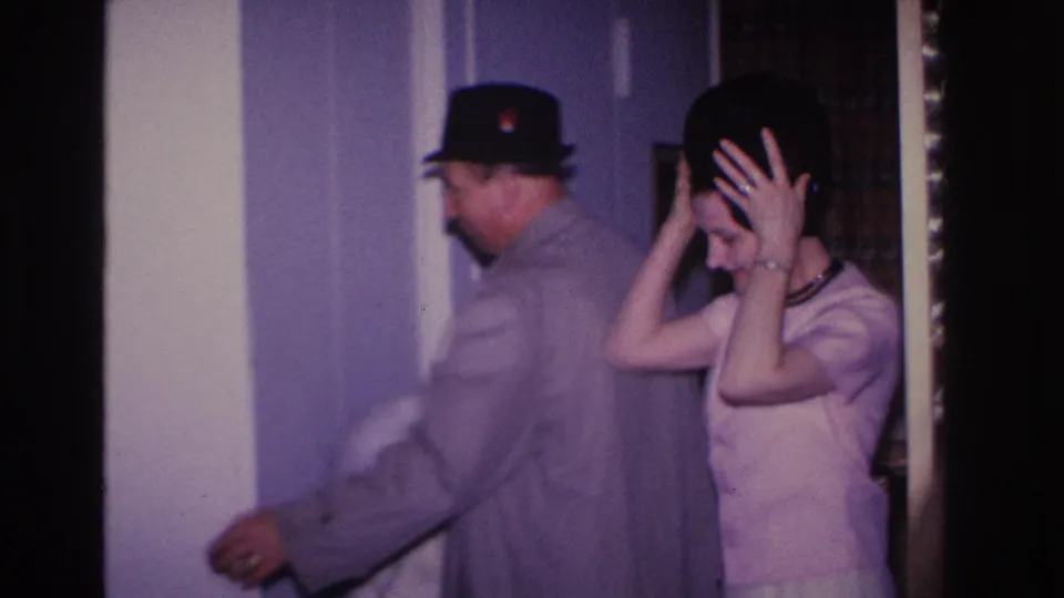 Man in gray overcoat and dark brim hat assists woman in pink dress who raises both hands to adjust a dark hat, residential interior circa 1966.
