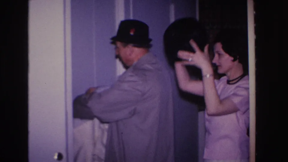 Man in gray overcoat and dark brim hat watches woman in pink dress settle a dark hat near a white paneled door, residential interior circa 1966 Hoboken, New Jersey.
