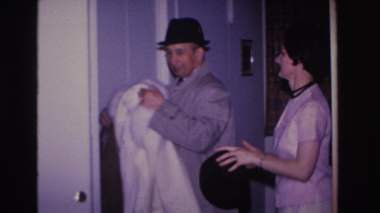 8mm home movie frame circa 1966, Hoboken NJ: man in black fedora holds white coat as woman in light pink shift dress reaches toward him near a purple-walled interior doorway.