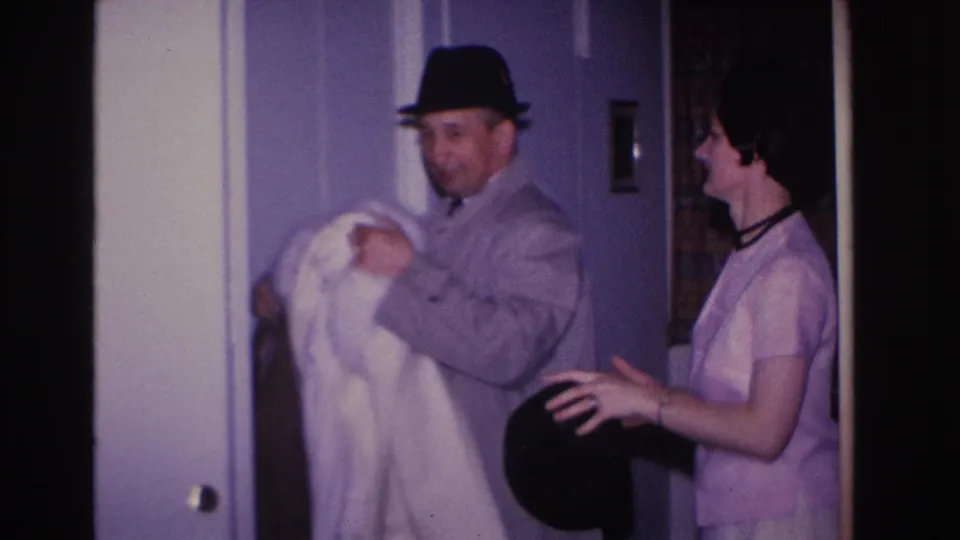 8mm home movie frame circa 1966, Hoboken NJ: man in black fedora holds white coat as woman in light pink shift dress reaches toward him near a purple-walled interior doorway.