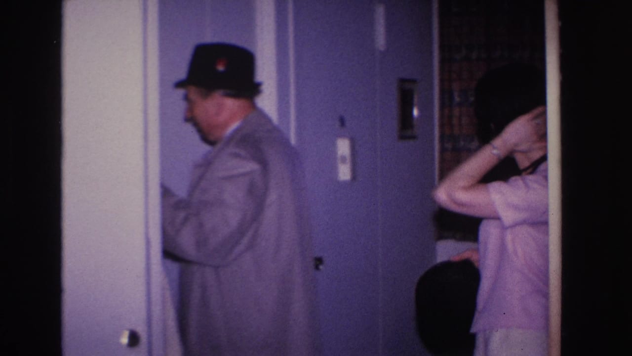 8mm home movie frame circa 1966, Hoboken NJ: man in overcoat and black fedora seen in profile near interior door as woman behind him raises her arm, possibly adjusting her hair.