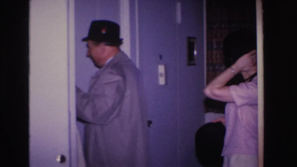 8mm home movie frame circa 1966, Hoboken NJ: man in overcoat and black fedora seen in profile near interior door as woman behind him raises her arm, possibly adjusting her hair.