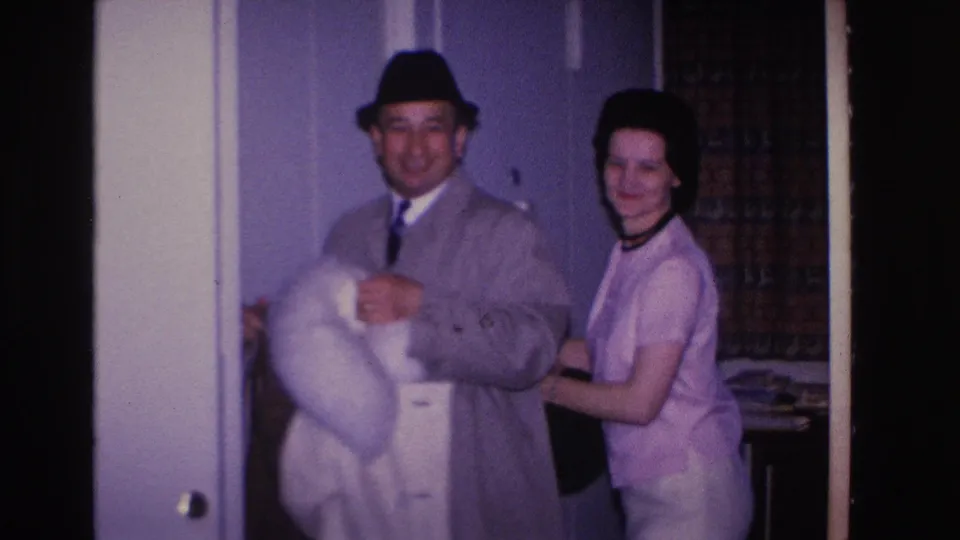 8mm home movie frame circa 1966, Hoboken NJ: man and woman smiling together indoors; he holds a white coat and wears a black fedora and blue tie, she wears a light pink dress and dark choker necklace.