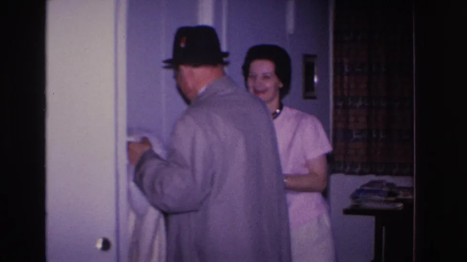 8mm home movie frame circa 1966, Hoboken NJ: man in overcoat seen from behind as woman in light pink dress smiles beside him near interior doorway with purple walls and patterned curtain.