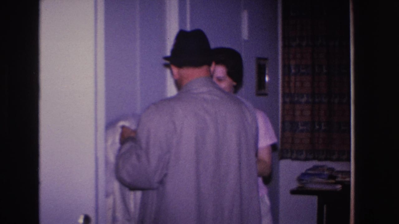 Man in black fedora and gray overcoat handles a white fur coat near an interior doorframe; woman in light short-sleeved dress stands alongside in a home interior, circa 1966.