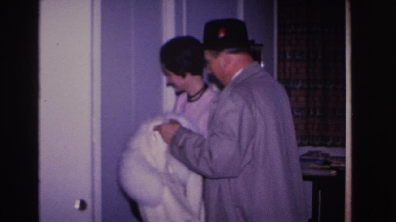 Man in black fedora and gray overcoat holds a white fur coat; woman with dark hair smiles warmly beside him in what appears to be a 1966 Hoboken home interior.