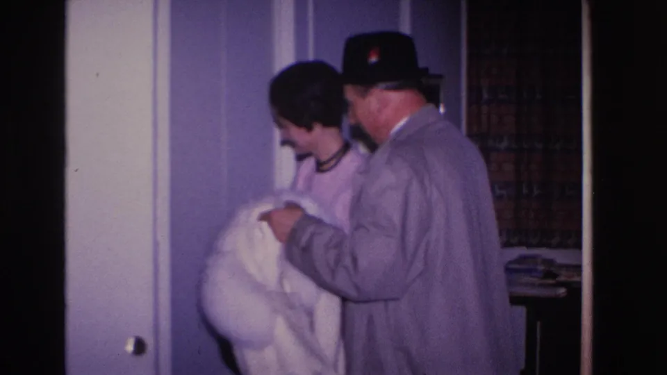 Man in black fedora and gray overcoat holds a white fur coat; woman with dark hair smiles warmly beside him in what appears to be a 1966 Hoboken home interior.