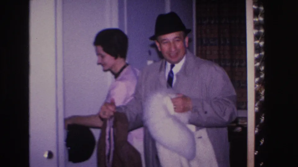 Older man in black fedora, gray overcoat, and blue tie smiles broadly holding a white fur coat; woman smiles beside him; holiday tinsel visible at frame edge, circa 1966 home interior.