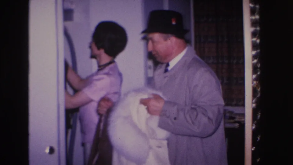 Man in black fedora and gray overcoat and woman with dark hair examine a white fur coat near a doorframe; holiday tinsel at right edge suggests a winter occasion, circa 1966 home interior.