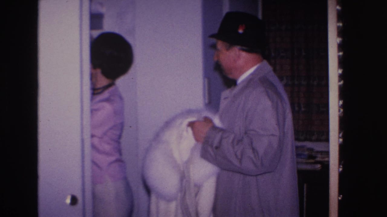 1966 8mm home movie: man in black fedora holds white fur coat while woman in pink dress and dark choker stands nearby in a home interior.