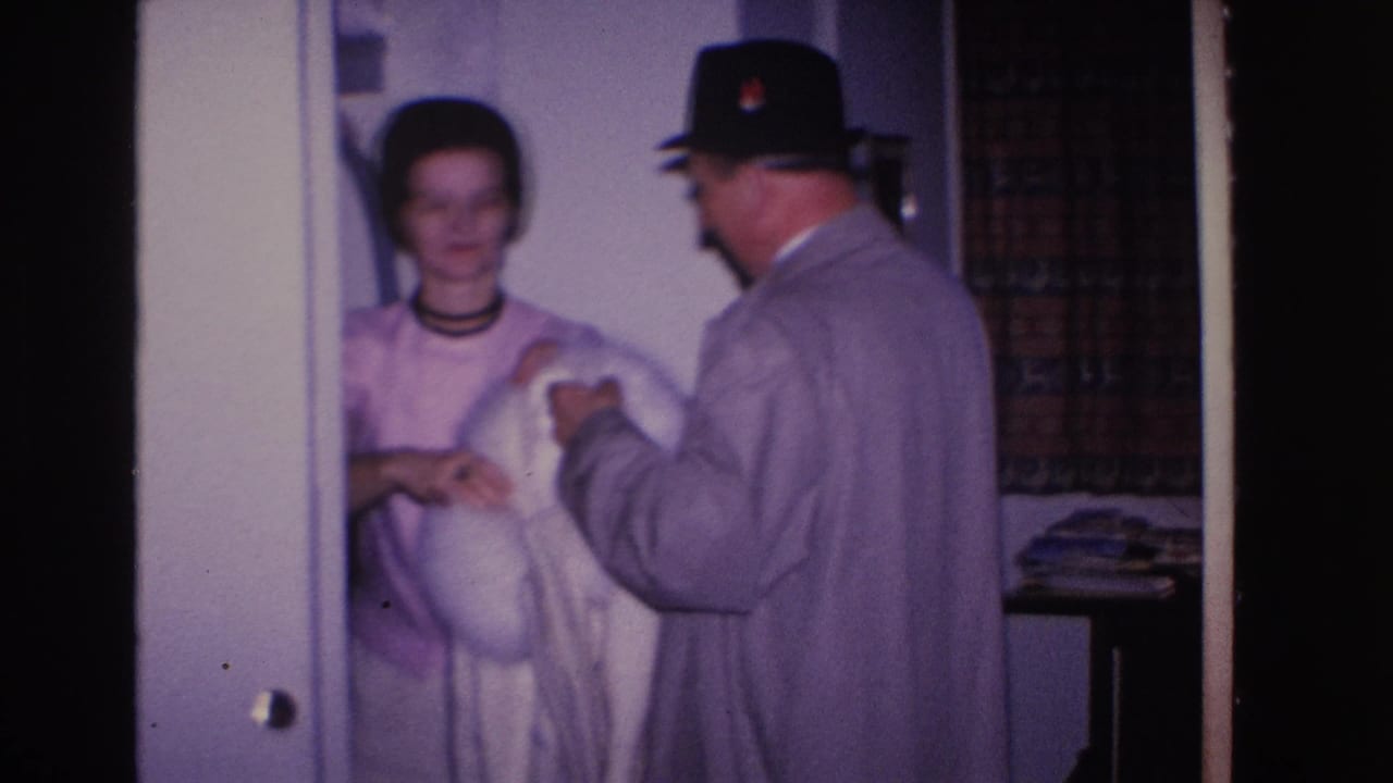 Woman in pink dress smiling as man in black fedora and gray coat holds white fur coat toward her.