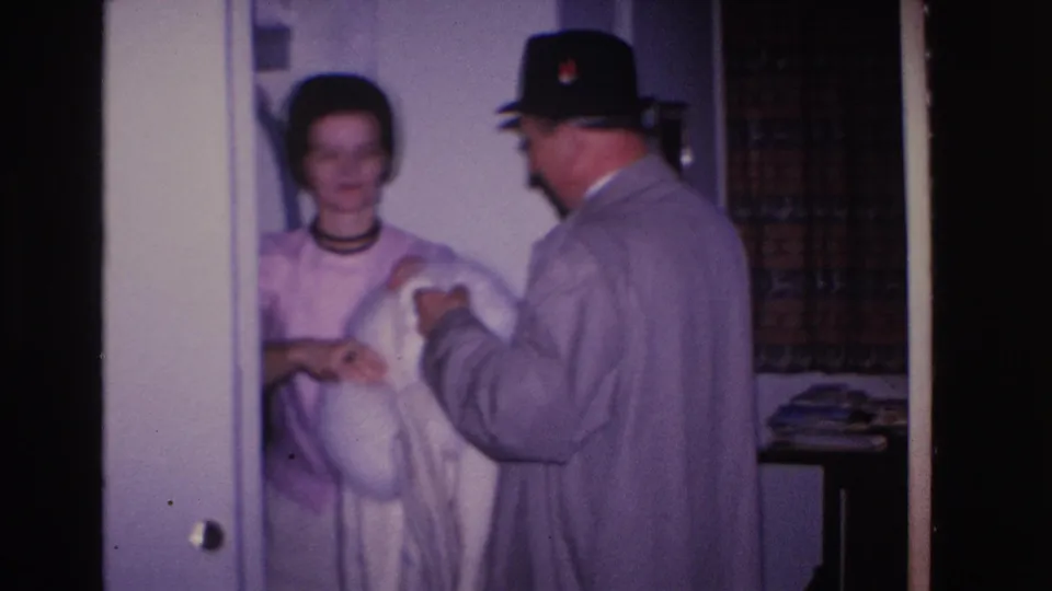 1966 8mm home movie: woman in pink dress with dark choker smiles as man in black fedora presents a white fur coat in a residential interior.