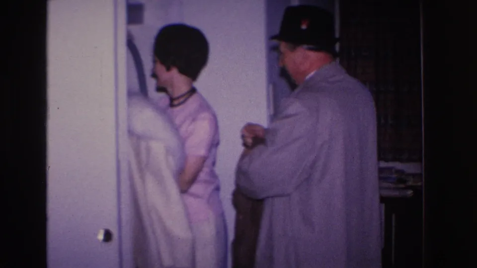 1966 8mm home movie: man in black hat and gray coat assists woman in pink dress and dark choker into a white fur coat near a residential doorway.
