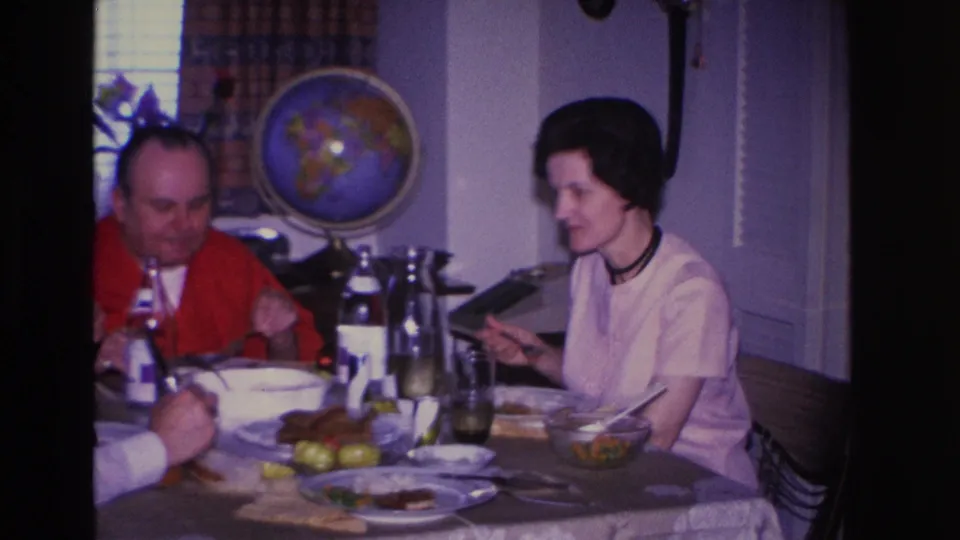 1966 8mm home movie: man in red shirt and woman in light top with dark choker dine at a table with food and bottles; a globe sits on a shelf in the background.