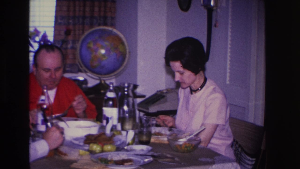 Man in red cardigan and woman in white blouse sharing a meal at a dining table in a mid-1960s American home interior; decorative globe visible on wall.
