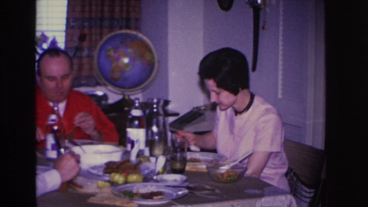 Man in red cardigan and woman in white blouse eating lunch in a 1966 home interior; a typewriter sits at the table edge with a globe on the wall behind.