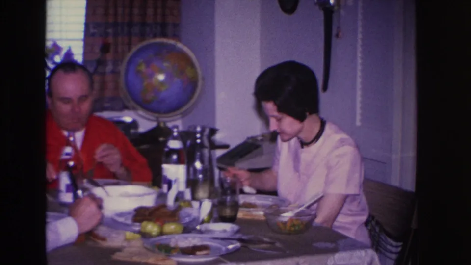 Man in red cardigan and woman in white blouse eating lunch in a 1966 home interior; a typewriter sits at the table edge with a globe on the wall behind.
