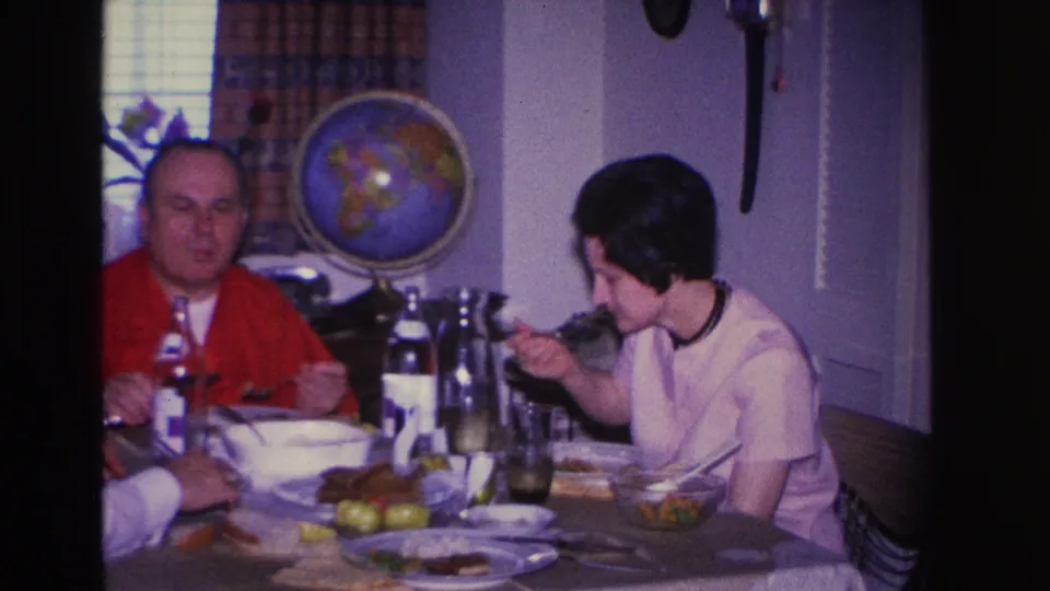 Woman in white blouse eating from a fork, man in red cardigan beside her; candid mid-meal moment in a 1960s domestic dining room with globe decor.
