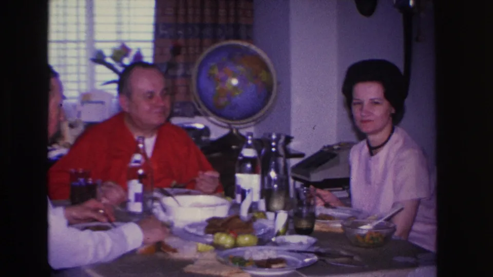 Woman in white blouse glances toward camera while man in red cardigan eats; plaid window curtains and decorative globe visible in this 1966 home-movie frame.