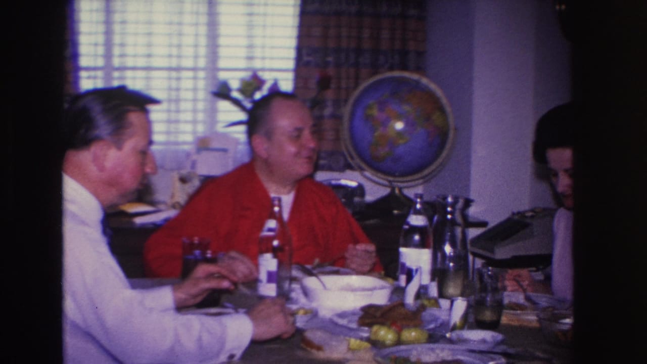 Two men at a set dining table in a 1966 domestic interior, with a woman partly in frame at right and a decorative globe and venetian-blind window visible behind them.