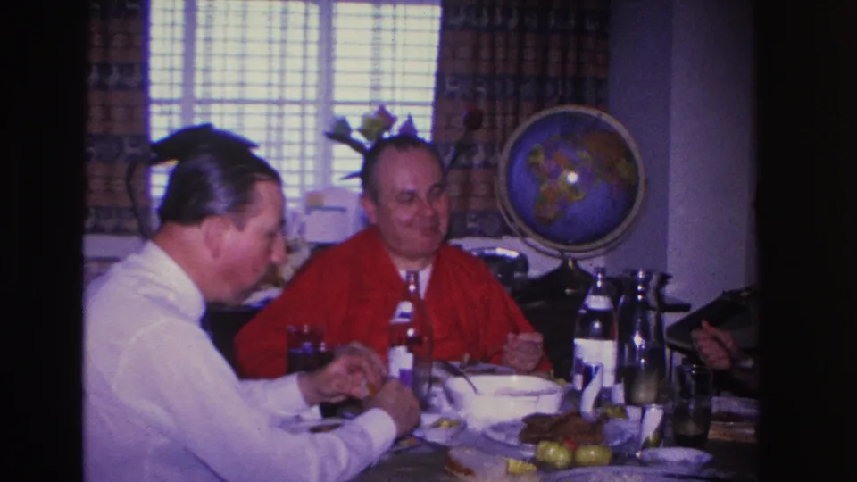 Two men eating at a domestic dining table in a 1966 home interior, globe on stand and venetian-blind window in background.