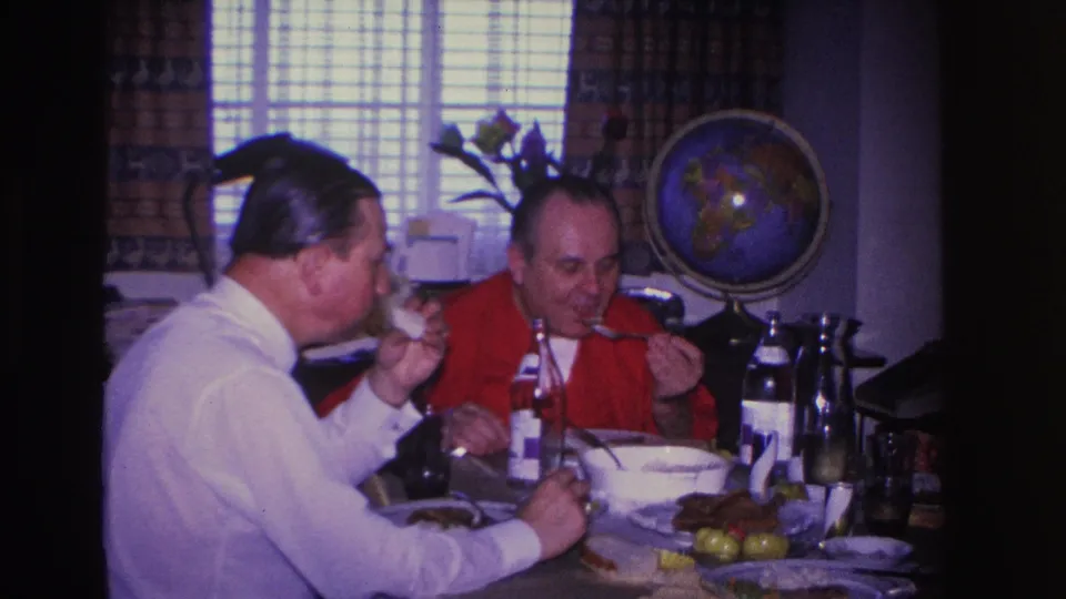 Two men mid-meal at a domestic dining table in a 1966 home interior, with a globe on a stand and venetian blinds visible behind them.