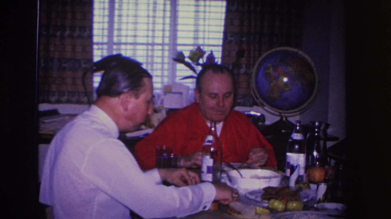 Two adult men at a home dinner table, circa 1966; globe on stand and window blinds visible in background behind them.