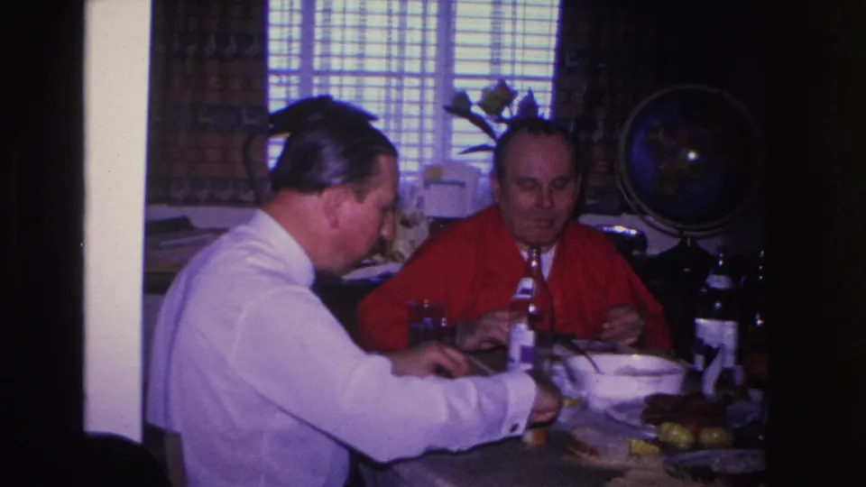 Two men at a domestic dinner table, circa 1966, Hoboken New Jersey area; bookcase visible at left edge, globe on stand and window behind them.