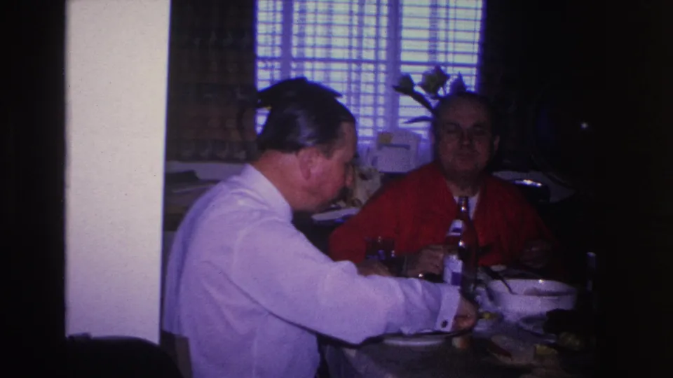Two adult men sharing a meal at home, circa 1966; man in white shirt leans toward table, globe on stand and vase with flowers visible in background.
