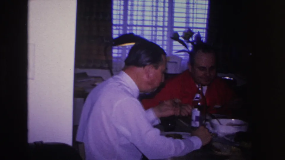 Two adult men eating from bowls at a private home dining table, Hoboken, New Jersey, circa 1966; 8mm footage with blue-shifted background light.