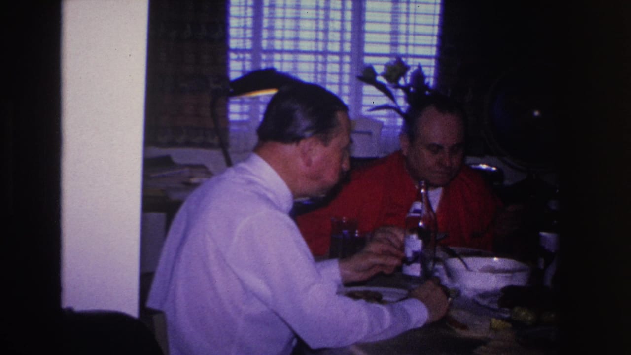 Two men continue eating at a dining table; venetian blinds and a plant are visible in the background.