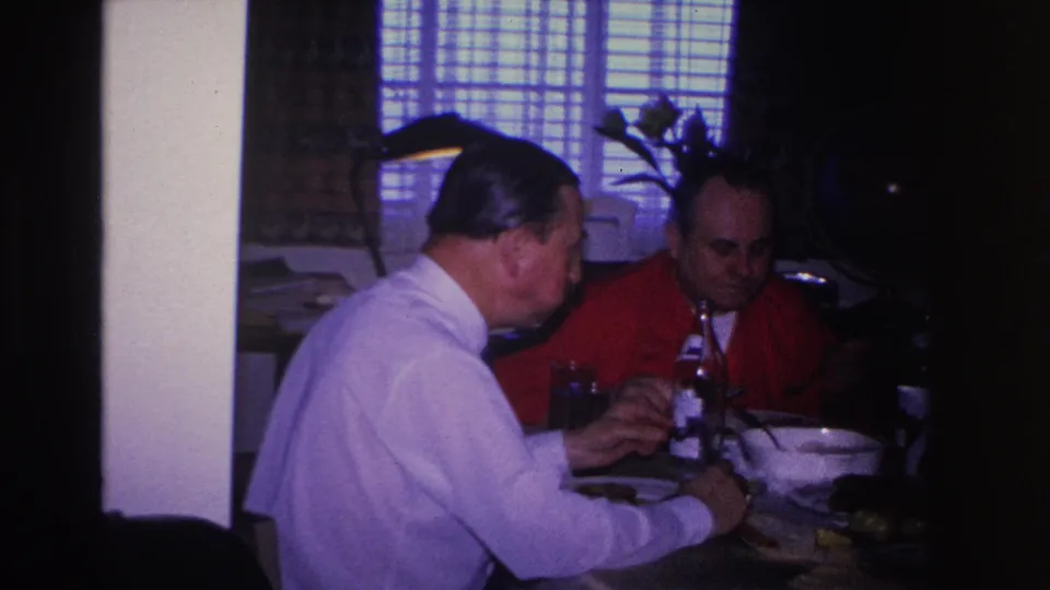 Two adult men at a mid-1960s dinner table in a private home; 8mm film with characteristic color shift and edge vignetting.
