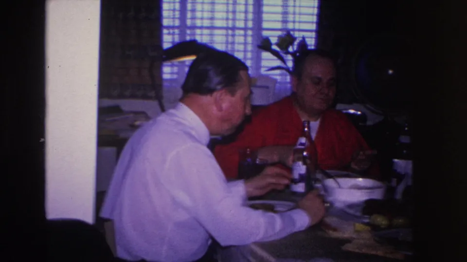 Slightly wider view of two adult men dining; man in red shirt partially faces camera; 8mm home movie, circa 1966 Hoboken, New Jersey.