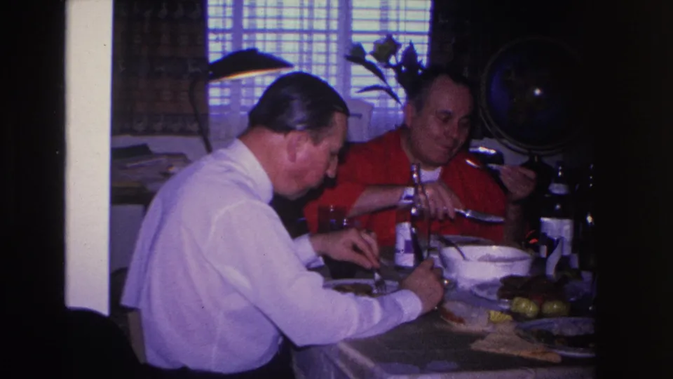 Two adult men near the end of a meal at a private home dining table; table set with bowls, dark bottle, and food items; circa 1966 8mm footage.