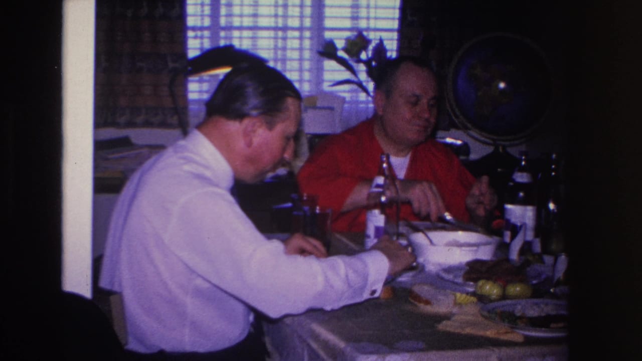 Two men share a meal at a set dining table in what appears to be a 1960s American home; a decorative globe is mounted on the back wall.