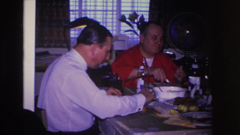 Two men share a meal at a set dining table in what appears to be a 1960s American home; a decorative globe is mounted on the back wall.