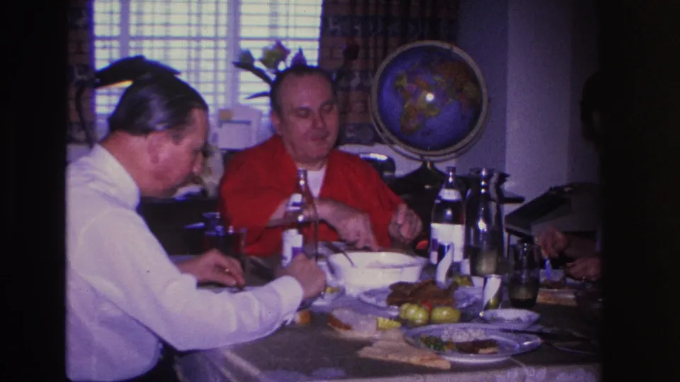 Two men eating lunch in a 1960s domestic interior; a large decorative globe and floral arrangement are clearly visible on the back wall.
