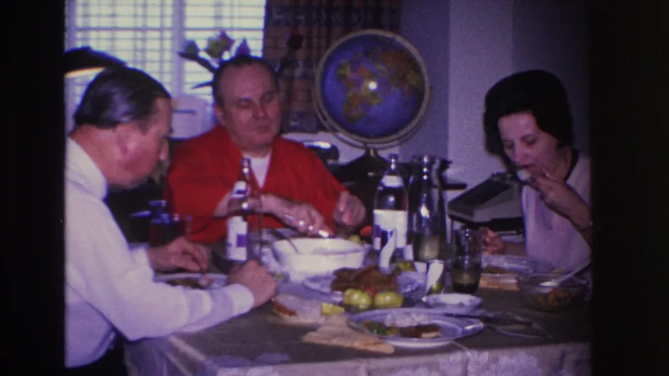 Three adults share a meal in a 1960s domestic interior, two men and a woman, with a decorative globe and window blinds in the background.