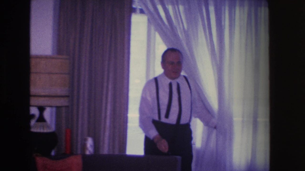 Middle-aged man emerging from sheer curtains in a domestic living room, circa 1966 Hoboken, NJ; table lamp and dark furniture visible at left.