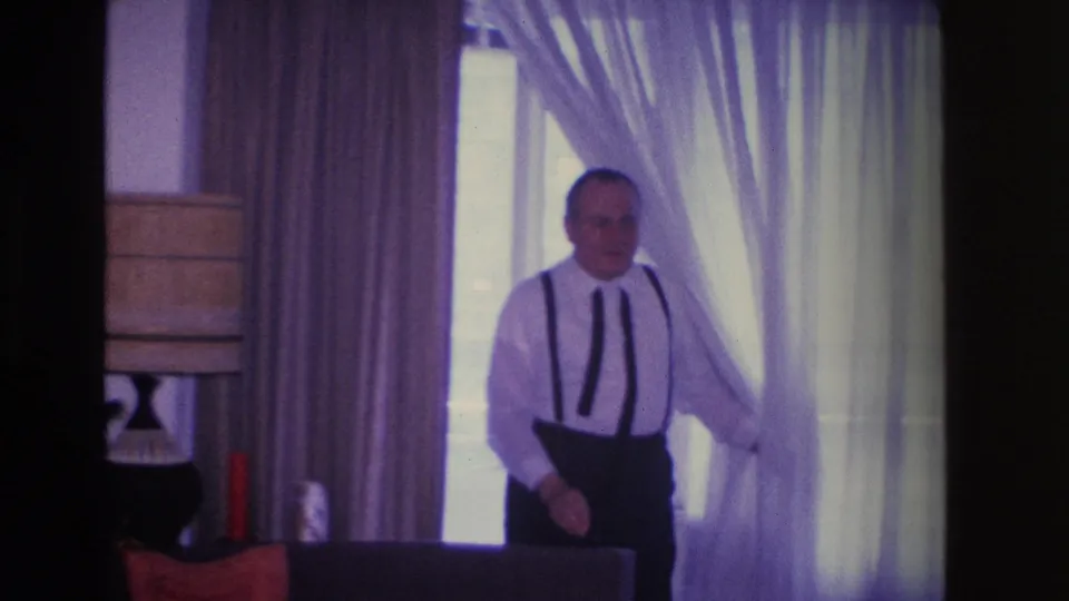Middle-aged man emerging from sheer curtains in a domestic living room, circa 1966 Hoboken, NJ; table lamp and dark furniture visible at left.