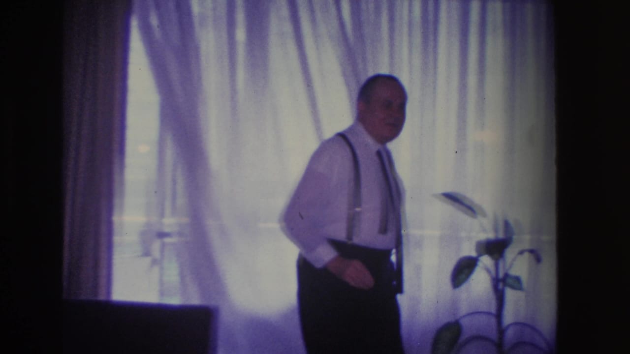 Man moving across a mid-century living room in front of sheer window curtains, circa 1966 Hoboken, NJ; potted plant on stand at right edge.