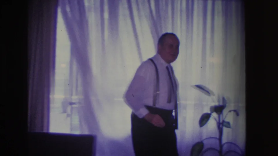 Man moving across a mid-century living room in front of sheer window curtains, circa 1966 Hoboken, NJ; potted plant on stand at right edge.