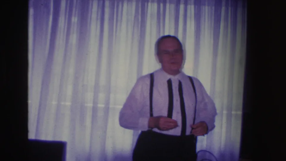 Man in 1960s dress attire advancing toward camera inside a domestic living room with sheer curtains behind, circa 1966 Hoboken, NJ.