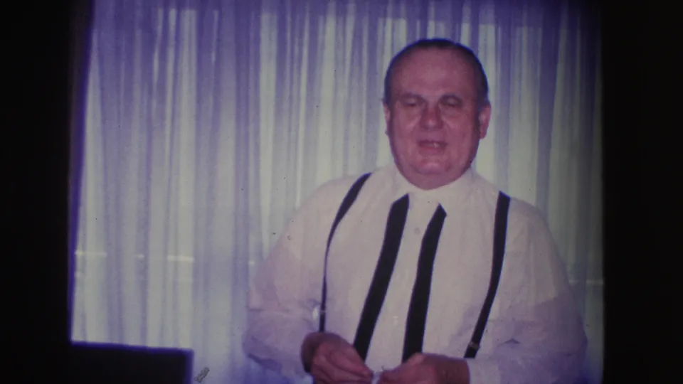Close waist-up shot of a man in 1960s dress attire facing camera with a slight expression; sheer curtains behind, domestic interior, circa 1966.
