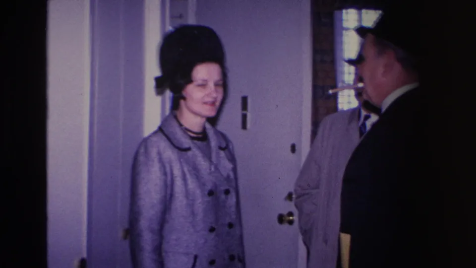 1966 home movie: woman in grey double-breasted coat and tall black hat at a residential doorway, man in dark suit at frame edge, Hoboken, New Jersey.